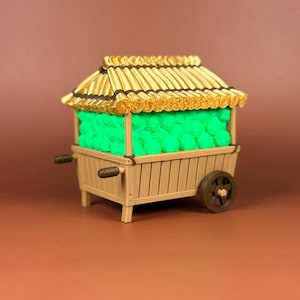 Peut inclure: Un chariot en bois miniature avec un toit de chaume, rempli d'objets vert vif. Le chariot a une roue et des poignées marron, et le toit est de couleur jaune doré. Le chariot est posé sur un fond marron.