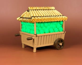 May include: A miniature wooden cart with a thatched roof, filled with bright green objects. The cart has a brown wheel and handles, and the roof is a golden yellow colour. The cart is set against a brown background.