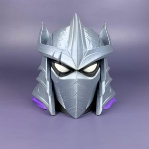 May include: A silver-colored 3D printed helmet featuring a pointed crest, sharp angular side panels, and a face mask covering the lower portion. The helmet has white eye openings and a visible purple accent piece at the base of the neck area.