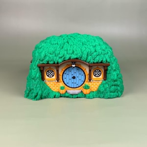 May include: A miniature hobbit-style house figurine featuring a bright blue circular door, yellow brick-patterned walls, and two small round windows. The structure is encased in a textured, vibrant green mound resembling a grassy hill.