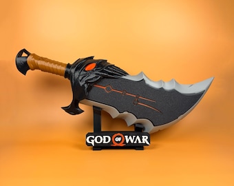 May include: A replica God of War blade in black and grey, featuring a brown handle and orange detailing. The blade is displayed on a black stand with the game's logo.