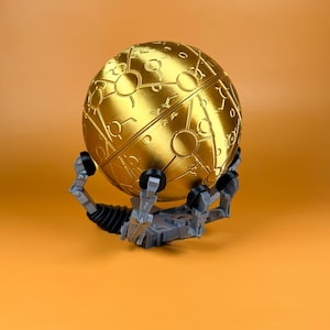 May include: A metallic gold spherical object featuring engraved circular and linear patterns across its surface. The sphere is held in place by a gray mechanical stand with articulated arms and black circular joints, creating a complex display.