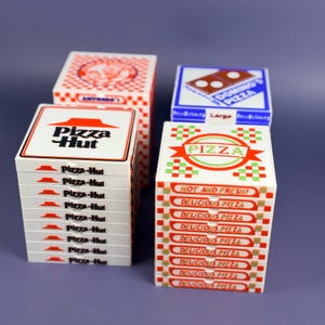 Può includere: Una collezione di scatole per pizza in miniatura di varie catene di pizzerie, tra cui Pizza Hut e Domino's. Le scatole sono bianche, rosse e blu, con loghi e testi come "Delicious Pizza" e "Hot and Fresh!" stampati.
