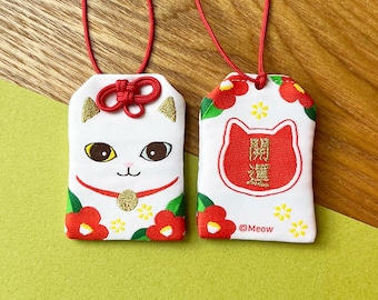 Könnte beinhalten: Zwei weiße japanische Glücksbringer-Katzen mit roten Kordeln. Ein Anhänger zeigt ein Katzen-Gesicht mit goldenen Akzenten und roter Schleife. Der andere Anhänger hat ein rotes Katzen-Design mit japanischen Schriftzeichen und dem Text "@Meow".