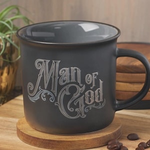 May include: A black ceramic mug with the text "Man of God" in a decorative font. The mug sits on a wooden coaster, with a stack of coasters and coffee beans in the background. A small plant is also visible.