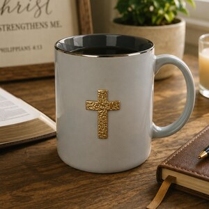 May include: A light gray ceramic mug with a gold cross emblem. The mug has a gray interior and a gold rim. The handle is light gray. The mug is on a wooden surface, with a framed text and an open book in the background.