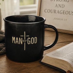 May include: A black ceramic mug with the words "MAN of GOD" in a cream-colored font, with a cross design. The mug has a large handle and sits on a wooden surface. An open book and a framed sign are also visible.