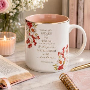 May include: White ceramic mug with a pink interior and gold trim, adorned with floral designs in red and white. The mug features the text "When she speaks, her words are wise & she gives instructions with kindness" and "Proverbs 31:26".