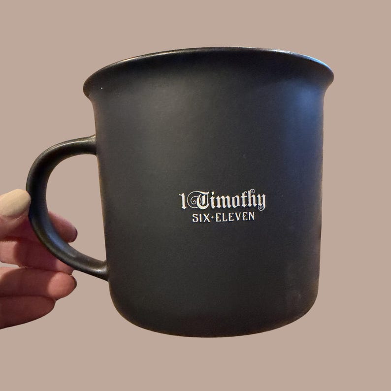May include: A black ceramic mug with a curved handle. The mug features the text "1 Timothy SIX ELEVEN" in white lettering. The mug is held by a person with a neutral background.