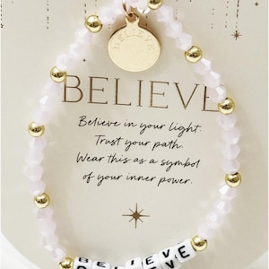 May include: A beaded bracelet with the word "BELIEVE" spelled out in white letter beads. The bracelet features pink faceted beads and gold-colored accents. The card behind the bracelet has the word "BELIEVE" in large letters and inspirational text.