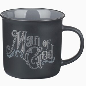 May include: A black ceramic mug with a gray interior and handle. The mug features the words "Man of God" in an ornate, silver-colored font. The design includes decorative flourishes.