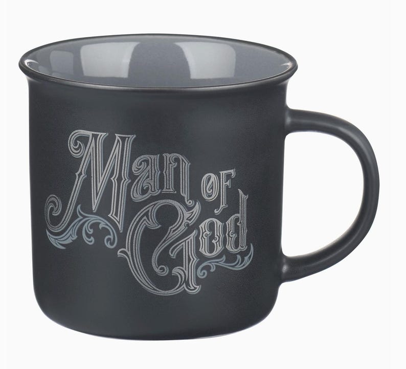 May include: A black ceramic mug with a gray interior and handle. The mug features the words "Man of God" in an ornate, silver-colored font. The design includes decorative flourishes.