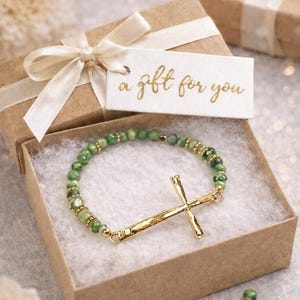 May include: A beaded bracelet with a gold cross charm rests in a small gift box. The bracelet features green beads and gold accents. A gift tag with the words "a gift for you" is attached to the box with a cream-colored ribbon.