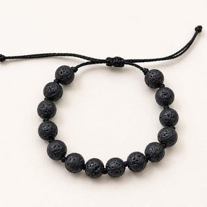 May include: A black bracelet featuring a series of round, porous lava stone beads strung on a thin black cord. The bracelet includes an adjustable sliding knot closure with two loose cord ends, each finished with a small knot at the very tip.