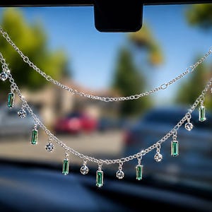 May include: A silver-toned double-layered chain car charm featuring alternating dangling emerald green rectangular crystals and clear round faceted stones. The delicate metal chains are suspended from a central point, creating a tiered effect.