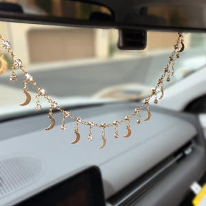 May include: A gold-toned chain hanging from a rearview mirror, featuring alternating dangling crescent moon charms and small clear faceted crystals. The delicate metal chain links connect each decorative element, creating a shimmering, layered appearance across the width of the mirror.