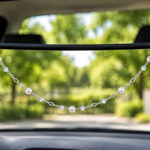 May include: A decorative car mirror hanging ornament featuring a silver-toned chain interspersed with white spherical beads, translucent light blue round beads, and small diamond-shaped metal links, suspended from a black rearview mirror.
