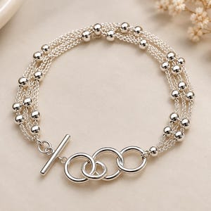 May include: A multi-strand silver bracelet featuring several delicate chains adorned with small, polished silver beads. The bracelet is secured with a toggle clasp consisting of a silver bar and three interlocking circular rings of varying sizes.