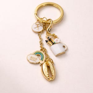 May include: Gold-coloured keychain with various charms. Charms include a white and gold initial pendant, a rainbow with "Back to School" text, a seashell, and a white mug with pencils and a flower. The keychain is set against a white backdrop.