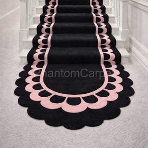 May include: Black stair runner with a scalloped pink border. The runner covers the center of the stairs, with a matching design at the bottom. The stairs have white risers and a wooden handrail.