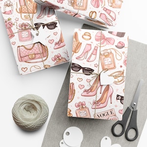 May include: Gift wrap featuring a repeating pattern of pink high heels, handbags, perfume bottles, sunglasses, and hearts on a white background. The wrapped gifts are tied with twine and adorned with star-shaped tags. A spool of twine and scissors are also present.