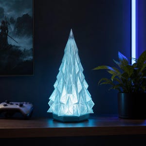 May include: A blue, geometric Christmas tree-shaped lamp. The lamp has a faceted design and emits a soft, cool light. It is placed on a wooden surface, with a video game controller and a potted plant in the background. The lamp is a decorative item.