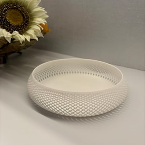 May include: A white, round decorative bowl with a lattice design. The bowl has a shallow interior and a textured exterior. The bowl is on a white surface. A sunflower is in the background.