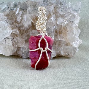 May include: A raw, deep red ruby pendant wrapped in silver-colored wire. The wire is intricately woven at the top and forms a decorative frame around the stone. The pendant is displayed against a backdrop of clear quartz crystals.