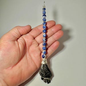 May include: A handmade witch's broom pendant. The pendant features a cluster of black stone at the bottom, with a series of blue and white beads strung above, connected by silver wire. The pendant is designed to hang.