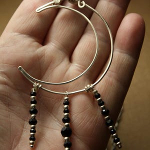 May include: A silver-toned crescent moon pendant necklace with three beaded dangles. The necklace features a detailed wire-wrapped design at the top and is adorned with black faceted beads and silver-colored beads.