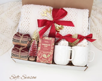 May include: A gift box filled with cozy items. It includes a cream-colored knit blanket, two white mugs, a box of chocolate chip cookies, and two pairs of patterned socks. The box is tied with a red ribbon and gold reindeer antlers.
