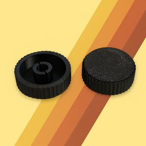 Puede incluir: Dos perillas negras y circulares con bordes estriados. Una perilla tiene un poste central con una ranura, mientras que la otra es un disco sólido. El fondo presenta rayas amarillas, naranjas y marrones.