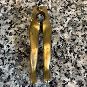 May include: A vintage brass nutcracker shaped like a pair of legs. The nutcracker is gold-colored and has a detailed design, including toes and a hinge at the top. The legs are slightly bent, suggesting a functional design.