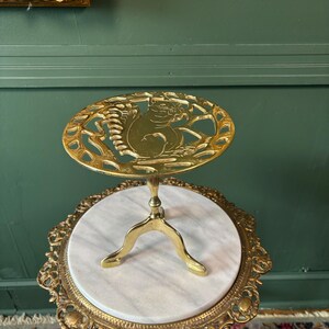 May include: A circular brass trivet featuring an openwork design of a squirrel sitting on a branch. The trivet is supported by a central pedestal base with three curved legs, all crafted from polished brass with a metallic gold-toned finish.
