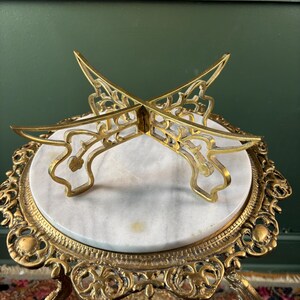 May include: A gold-toned metal display stand with an X-shaped top featuring intricate cut-out patterns. The stand rests on a circular white marble base, which is supported by a decorative gold-toned metal frame with ornate, curved legs and detailing.