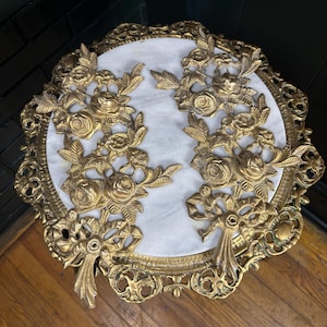 May include: A round table featuring a white marble top and an ornate gold-colored metal frame. The surface is decorated with two symmetrical gold-colored metal floral appliqués, each depicting roses, leaves, and ribbons, arranged vertically.