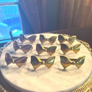 May include: A set of eight gold-toned metal napkin rings, each featuring a polished bow-shaped design. The napkin rings are arranged in a grid pattern, showcasing a reflective metallic finish and a structured, symmetrical bow silhouette.