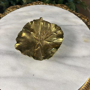 May include: A small, gold-toned metal dish shaped like a textured leaf with a central stem detail. The dish features a polished, reflective surface with organic, irregular edges and a concave center, mimicking the appearance of a natural plant leaf.