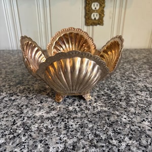 May include: A silver-toned metal bowl shaped like a large scallop shell with a scalloped rim and three small feet. The bowl features a ribbed, fluted design radiating from the center, with a polished metallic finish reflecting light across its surface.