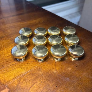 May include: A set of twelve polished gold-tone metal cabinet knobs arranged in a grid. Each knob features a smooth, rounded, mushroom-shaped head with a reflective surface and a short, cylindrical base designed for attachment to furniture or doors.