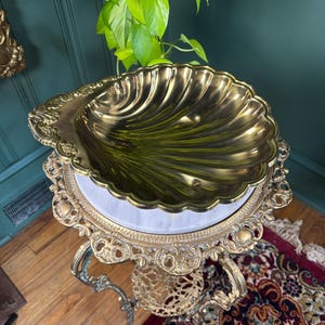 May include: A polished brass bowl shaped like a large scallop shell with a scalloped edge and radial ridges. The bowl rests on a decorative brass pedestal stand featuring intricate scrollwork, a circular marble top, and a small lower shelf.