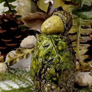 May include: A ceramic acorn figurine with a green leafy base. The acorn is painted in shades of brown and green, with a detailed texture. The base is decorated with a leafy pattern and has a rustic look.