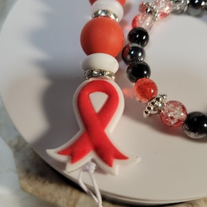 Puede incluir: Un juego de llavero y pulsera. El llavero presenta cuentas rojas y blancas, con un dije de cinta roja. La pulsera tiene cuentas negras, rojas y transparentes. Ambos artículos tienen detalles plateados.
