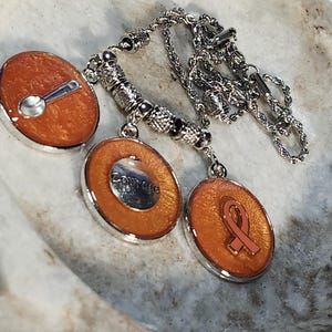 May include: A silver-toned necklace featuring three circular pendants with a shimmering orange resin finish. One pendant displays a small silver spoon, another features a silver disc with the word Courage, and the third shows a peach-colored ribbon.