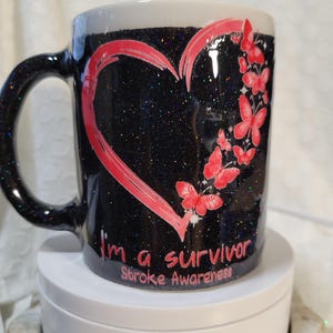 May include: Black glitter mug with a red heart and butterflies design. The mug has the text "I'm a survivor Stroke Awareness". The handle and rim are black. The mug is on a white surface.