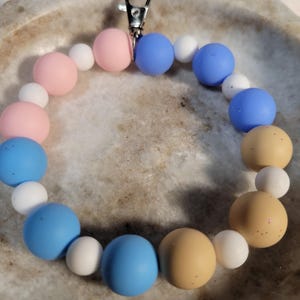 May include: A beaded keychain wristlet featuring a variety of colorful beads. The beads are in shades of pink, white, blue, and tan. A silver-colored clasp is attached to the wristlet.