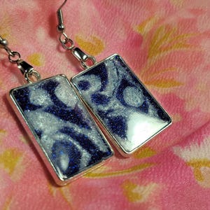 May include: Silver-toned rectangular earrings with a swirling design of navy blue and silver glitter set in resin. The earrings have a hook closure and are displayed on a pink floral background. These earrings are a unique accessory.