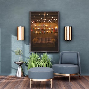 Puede incluir: Una obra de arte enmarcada que presenta una escena de sala de estar con un sofá, luces de cadena y una exhibición del alfabeto. La habitación está decorada con una silla azul, una otomana y apliques de pared. También hay una pequeña planta y una mesa auxiliar.