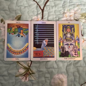Könnte beinhalten: Drei Tarotkarten, die horizontal angeordnet sind. Die erste Karte ist die Zehn der Kelche, die auf dem Kopf steht. Die zweite Karte ist die Neun der Schwerter, die eine Person auf einem Bett zeigt. Die dritte Karte ist Der Wagen mit einer Figur.