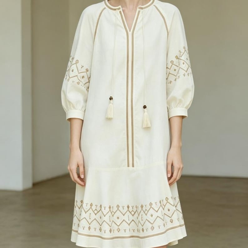 May include: Cream-colored dress with embroidered details. The dress features a V-neck, three-quarter sleeves, and a flared hem. Brown trim and tassels accent the neckline and front. Geometric embroidery adorns the sleeves and hemline, creating a bohemian aesthetic.